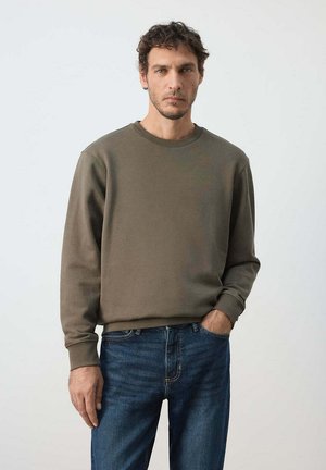 Hombre con cabello corto y rizado, vestido con sudadera de color verde oliva y vaqueros azules, de pie con una mano en el bolsillo frente a un fondo liso.