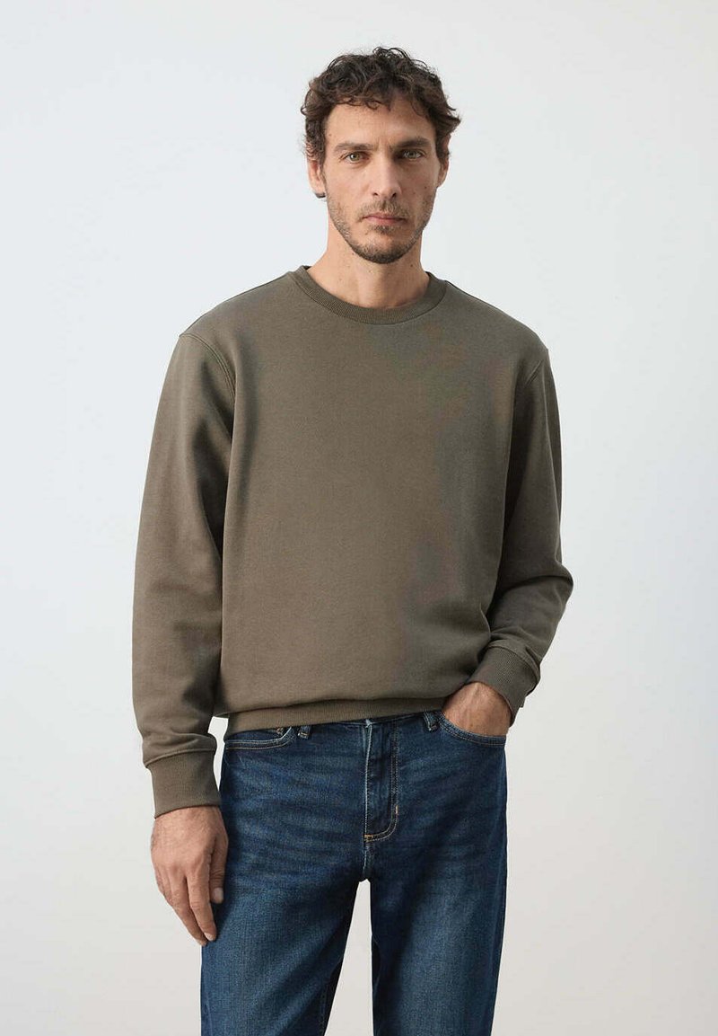 Homme aux cheveux courts et bouclés, portant un sweat-shirt vert olive et un jean bleu, debout avec une main dans la poche devant un fond uni.