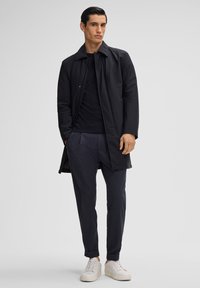 Schwarzer leichter Mantel mit spitzem Kragen, über einem dunklen Pullover getragen, kombiniert mit dunklen Hosen und weißen Sneakers. Einfaches, sauberes Design.