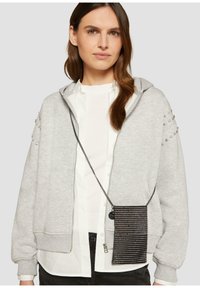 Szara bluza z zamkiem błyskawicznym z ozdobnymi ramionami, nałożona na białą koszulę, w połączeniu z czarną torebką na ramię pokrytą cekinami na srebrnym łańcuszku.