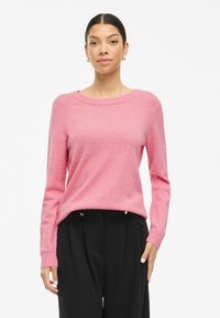 Pink knit sweater with a round neckline, long sleeves, ribbed cuffs, and a relaxed fit, worn over black trousers. Smooth texture.
