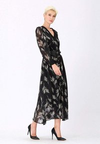 Femme blonde dans une robe midi noire transparente à manches longues avec un imprimé plumes, ceinture noire et chaussures à talons hauts noires se tient contre un fond blanc.