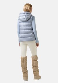 Hellblauer gepolsterter Weste mit Kapuze, die über gesteppten Bereichen verfügt. Kombiniert mit beigen gepolsterten Beinstulpen und hellfarbigen Hosen.