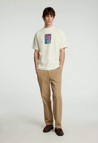 Un giovane uomo sta in piedi con le mani nelle tasche, indossando una t-shirt bianca con grafica, pantaloni beige e scarpe marroni, su uno sfondo chiaro e semplice.