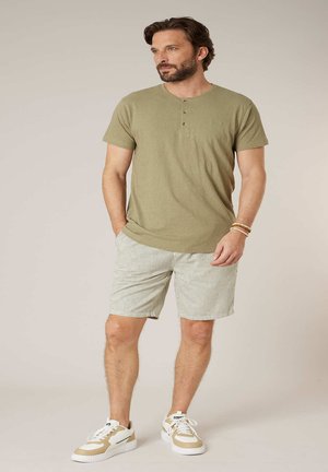 Man met baard die een olijfgroen henleyshirt draagt, lichte gestreepte korte broek, witte en beige sneakers, en armbanden, staat met zijn handen in zijn zakken.