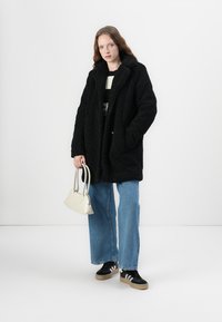 Black, fluffy, oversized coat with a collar, paired with a striped top, light blue jeans, black sneakers, and a small white handbag.