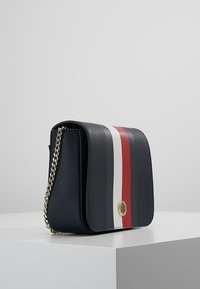 Bolso de hombro de cuero azul marino con forma rectangular plana, que presenta rayas verticales blancas y rojas y un detalle del logotipo en dorado. Incluye cadena.