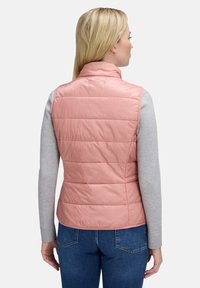 Jasnoróżowa, bezrękawnik, pikowany, z wysokim kołnierzem, teksturowana tkanina oraz szwy na plecach. Noszona na szarym długim rękawie i niebieskich dżinsach.