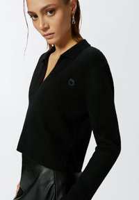 Crni pleteni pulover s rebrastim pojasom, ovratnikom i dugim rukavima. Ima mali izvezeni logo na lijevoj strani prsiju.