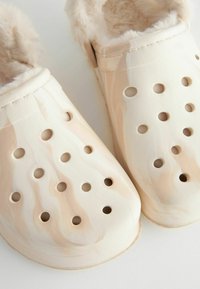 Zoccoli beige chiaro realizzati in materiale sintetico, dotati di una morbida fodera in pelliccia sintetica, numerosi fori di ventilazione e una superficie liscia e leggermente lucida.