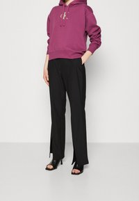 Purple hoodie with a logo, paired with black trousers featuring side slits. Model wears black heeled sandals. Simple design and comfortable fit.