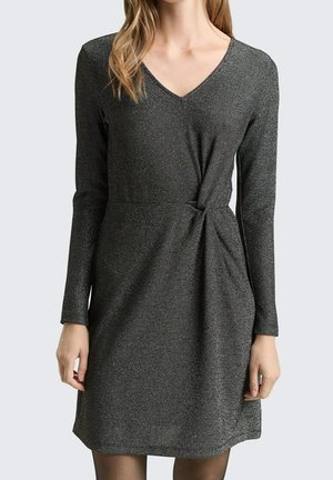 Kobieta ubrana w długą, ciemnoszarą sukienkę z połyskiem, z dekoltem w kształcie litery V i marszczeniem w talii, stojąca na tle jasnego, jednolitego tła.