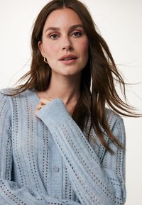 Ljusblå stickad kofta med vertikala ränder, öppen stickning och knäppning framtill. Har långa ärmar och en mjuk textur.