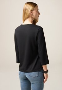 Frau mit langen blonden Haaren, die ein schwarzes Oberteil mit drei Viertelärmeln, eine blaue Jeans, lange hängende Ohrringe und ein schwarzes Perlenarmband trägt.