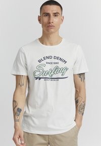 Bílý bavlněný tričko s kulatým výstřihem a kontrastním grafickým potiskem se slovem "Blend Denim Surfing Built in Blue." Krátké rukávy s viditelnými tetováním.