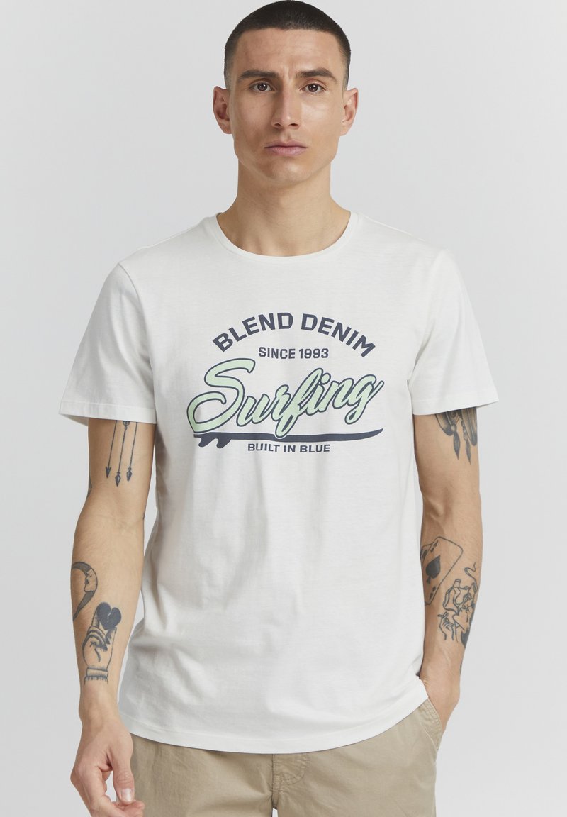 Bílý bavlněný tričko s kulatým výstřihem a kontrastním grafickým potiskem se slovem "Blend Denim Surfing Built in Blue." Krátké rukávy s viditelnými tetováním.