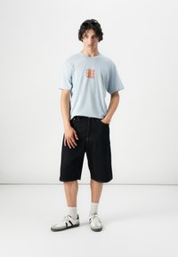 Camiseta de color azul claro con texto naranja "ALIMENTA TU MENTE"; pantalones cortos de mezclilla oscura; zapatos blancos con detalles negros; calcetines grises; ajuste casual.