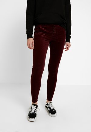 Osoba nosząca bordowe aksamitne spodnie o dopasowanym kroju z guzikami z przodu oraz czarno-białe trampki, stojąca na jednolitym białym tle.