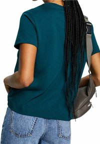 Person med långt flätat hår som bär en teal kortärmad skjorta och ljusblå jeans, hållande en grå axelväska.