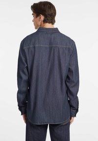 Tummansinisestä denimmistä valmistettu pitkähihaiseen paitaan, jossa on napillinen kaulus ja yksinäiset napit hihansuissa. Paita on suoralinjainen helma ja takana on ommeltu sauma.