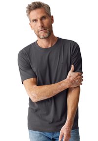 Graues kurzärmeliges T-Shirt aus weichem Stoff, mit rundem Ausschnitt und einem dezenten Logo auf der Brust. Modell trägt Jeans.