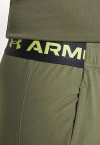 Primer plano de pantalones verde oliva con cintura elástica negra que presenta un logo parcial amarillo de Under Armour y texto.