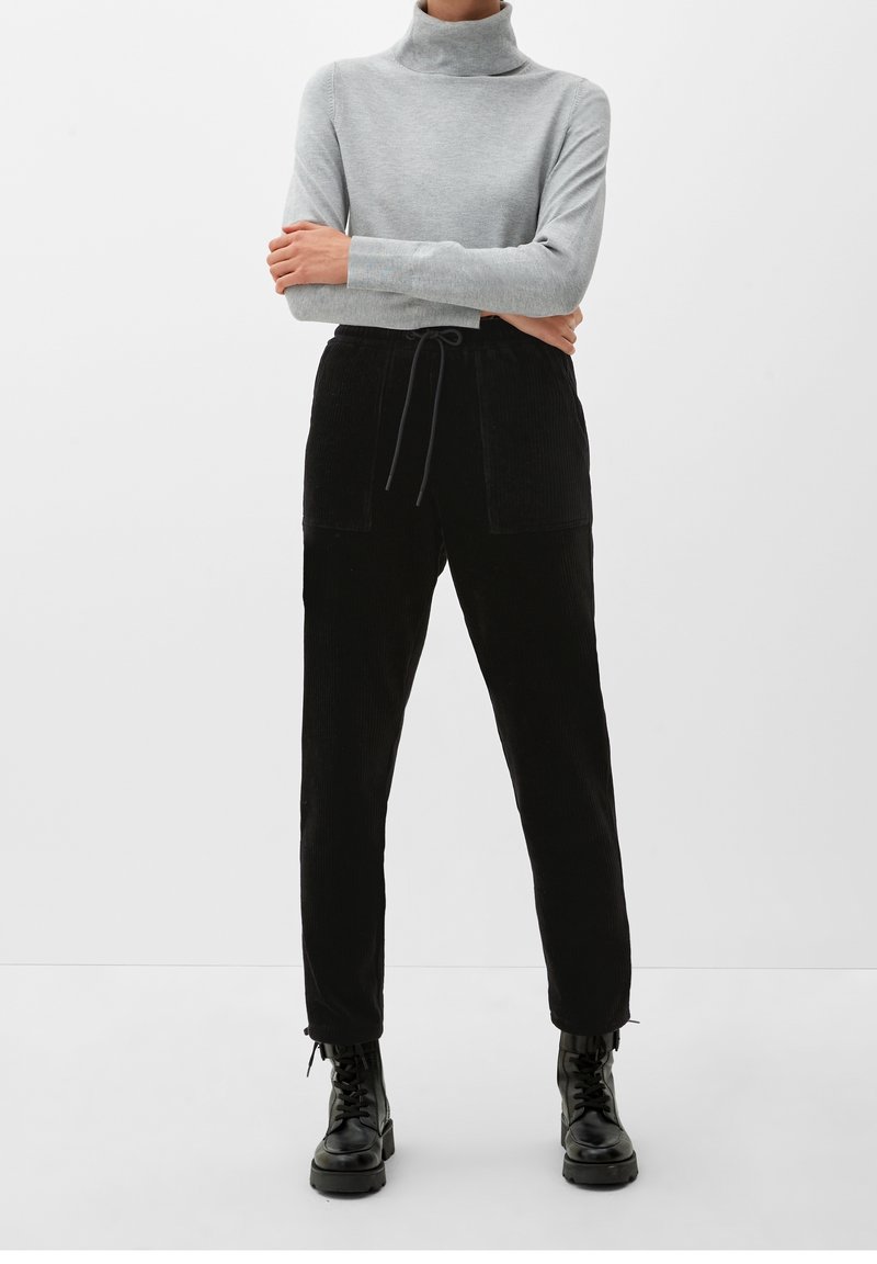Pull à col cheminée gris, style ajusté, associé à un pantalon en velours côtelé noir taille haute avec deux poches avant et un cordon de serrage ajustable. Bottes noires à lacets.