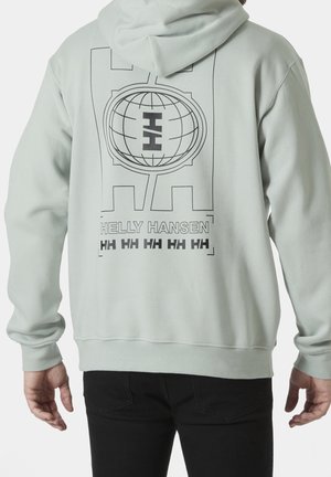 Osoba ubrana w jasnoszarą bluzę z kapturem Helly Hansen z nadrukiem globu i logo "HH" na plecach, w połączeniu z czarnymi spodniami.