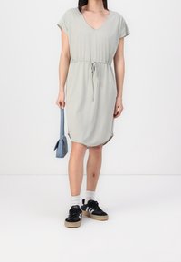 Hellgrünes, kurzärmliges Kleid mit V-Ausschnitt, tailliertem Bund und knie-langer Länge. Kombiniert mit schwarzen Sneakers und einer kleinen blauen Tasche.