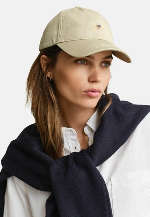 Femme portant une casquette beige avec un écusson brodé, une chemise blanche boutonnée et un pull bleu marine drapé sur les épaules, sur un fond uni.