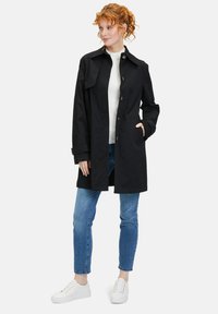 Schwarzer Trenchcoat mit Knopfverschluss, breitem Kragen, zwei Seitentaschen und geradem Schnitt, kombiniert mit blauen Jeans und weißen Sneakern.
