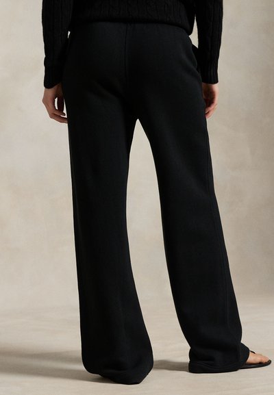 Person iført sorte vidde bukser og en sort kabelstrikket sweater, stående barfodet på en neutral baggrund.