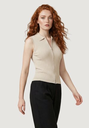 Donna con capelli rossi lunghi e ricci indossa un top smanicato beige a coste con cerniera e pantaloni neri, in piedi contro uno sfondo chiaro e semplice.