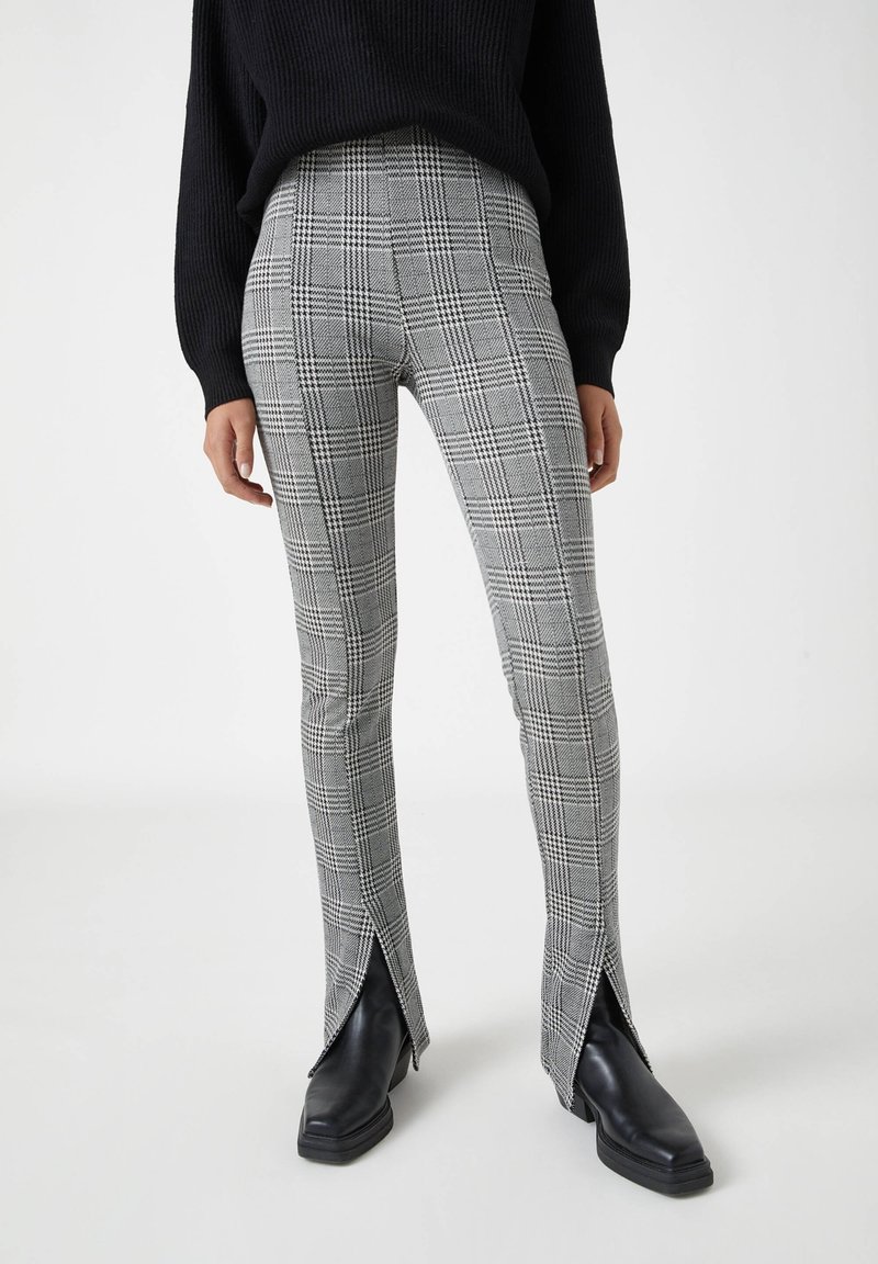 Femme portant un pull noir, des leggings gris à carreaux avec des fentes à l'avant des chevilles, et des bottes noires à bout carré, debout sur un sol blanc.