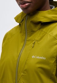 Κοντινό πλάνο προσώπου που φορά ένα πράσινο μπουφάν Columbia με φερμουάρ στο στήθος και στο εμπρόσθιο μέρος, εμφανίζοντας το λογότυπο της μάρκας στο στήθος.