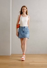 Modèle portant un débardeur blanc, une jupe en denim bleu et des sandales roses avec des accents oranges, portant un petit sac orange. Fond en parquet en bois.