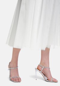 Sandales à talons hauts en argent avec de fines brides ornées de strass. Le talon est de type stiletto, complétant une jupe blanche transparente.