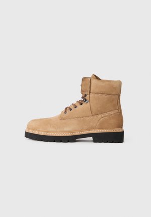 Beez tan suede pahklottud saapad ümarate varvastega, nööridega ette ja mustriliste nööpidega. Omab tekstuuriga krae ning musta kummist tallaga, millel on mustriga kumm.