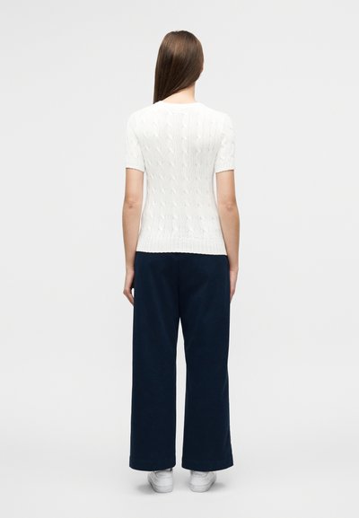 Λευκό πλεκτό πουλόβερ με κοντά μανίκια και στρογγυλή λαιμόκοψη, σε συνδυασμό με μπλε-σκούρο παντελόνι φαρδιάς γραμμής και λευκά αθλητικά παπούτσια.