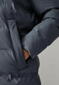Marineblaue Steppjacke mit einer glatten, gequilteten Textur, ausgestattet mit einer Seitentasche und einer lockeren Passform. Die Ärmel sind weit und gepolstert für zusätzliche Wärme.