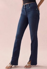 Jeans di lavaggio scuro con gamba svasata, caratterizzati da spaccature laterali e accessori in tono rame. Realizzati in denim, progettati per una vita aderente.