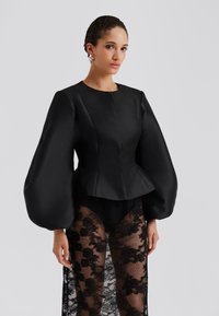 Μαύρο σατέν τοπ με πεπλούμ και φαρδιές, ογκώδεις μανίκες. Το κάτω μέρος διαθέτει διαφανές μαύρο δαντελένιο επικάλυμμα με λουλουδάτα σχέδια, διπλωμένο πάνω σε σκούρα εσώρουχα.