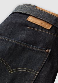 Jean en denim bleu foncé plié montrant la poche arrière, des coutures jaunes contrastantes et une étiquette de taille en cuir marron sur la ceinture.