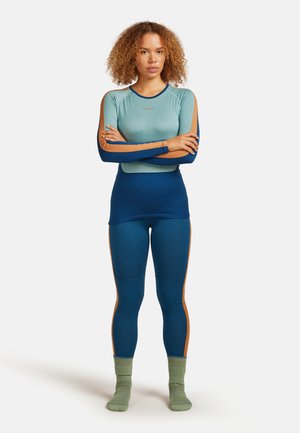 Donna con capelli ricci sta in piedi con le braccia incrociate, indossando un top a maniche lunghe blu e beige, leggings abbinati e calzini verdi su uno sfondo bianco.