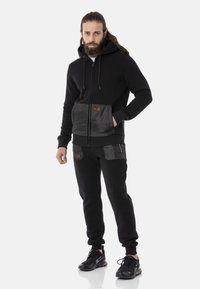 Schwarzes Hoodie mit Reißverschluss vorne, kontrastierenden Stofftaschen und Logopatches. Kombiniert mit schwarzen Jogginghosen, die beide eine lockere Passform haben.