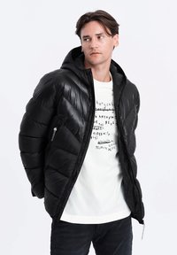 Schwarze Pufferjacke mit Kapuze, ausgestattet mit einer Reißverschlusstasche und einem rautenförmigen Steppmuster. Getragen über einem weißen T-Shirt mit Grafik.