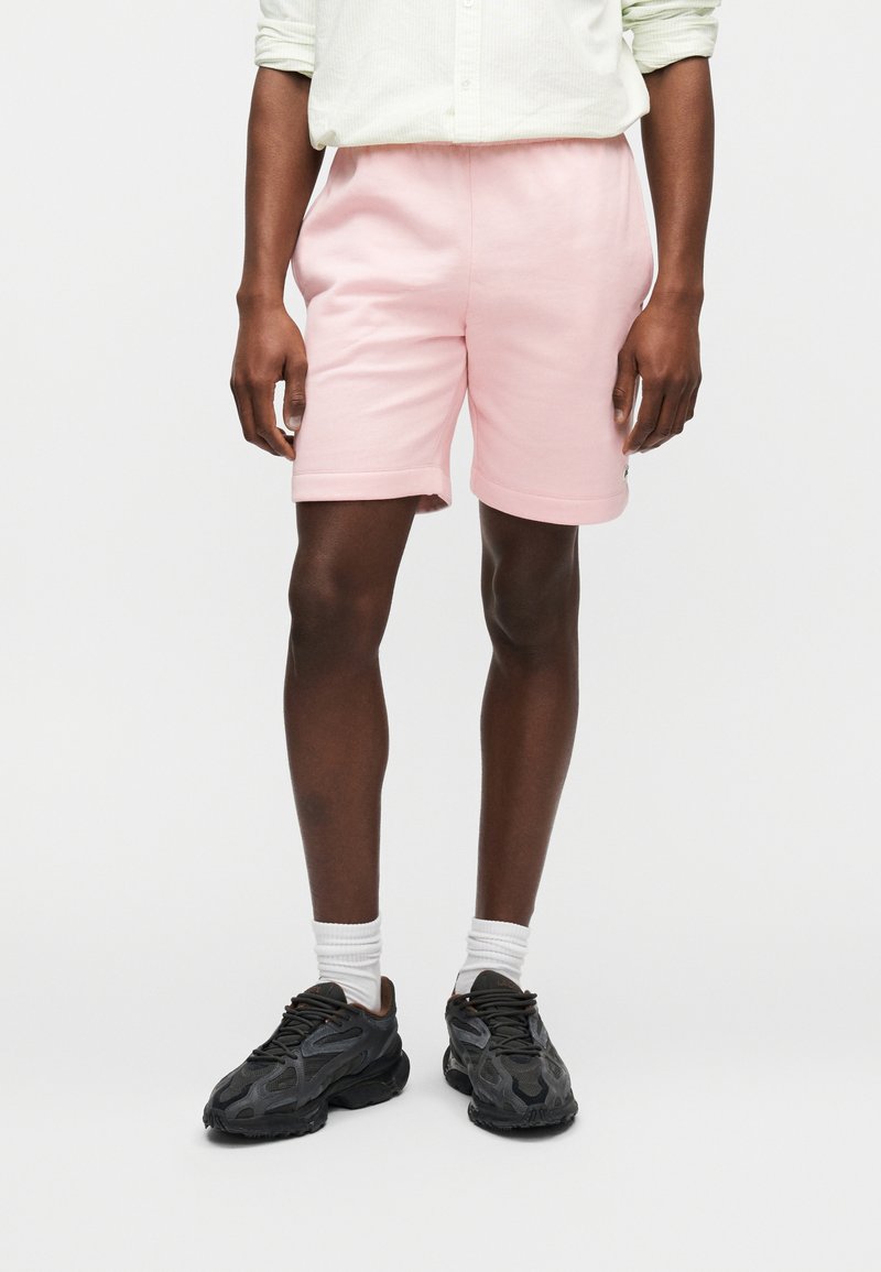 Άτομο που φοράει ροζ σορτς με διπλωμένες άκρες, λευκές κάλτσες, μαύρα αθλητικά παπούτσια και ανοιχτόχρωμο πουκάμισο, στέκεται μπροστά σε ένα απλό φόντο.