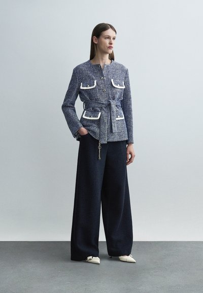 Γυναίκα με μπλε καρό σακάκι με ζώνη και τσέπες με λευκή περιγραφή, και φαρδιά μαύρη παντελόνα, στέκεται μπροστά σε απλό γκρι φόντο.