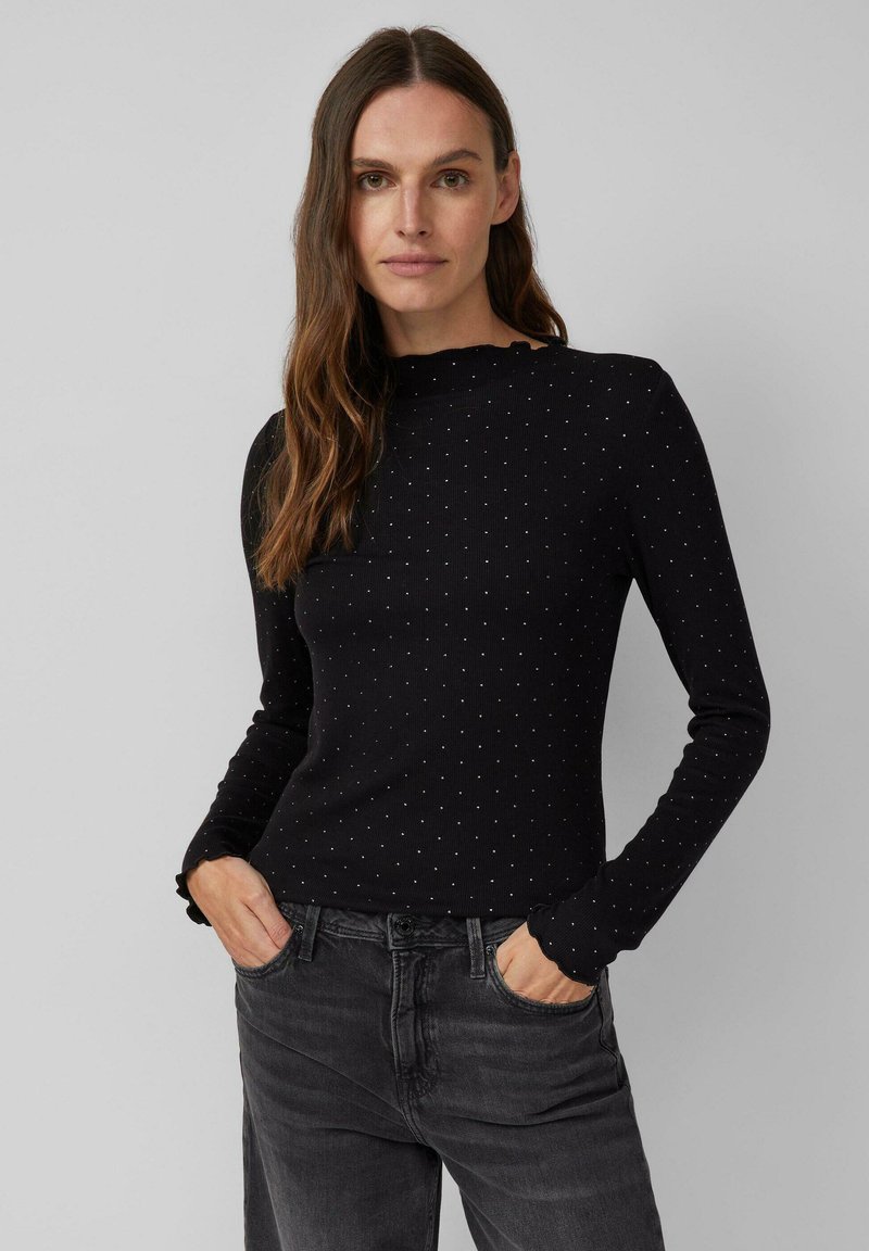 Zwarte lange mouwen top met een geribbelde textuur en witte stippen. Heeft een subtiele volants halslijn en een aansluitende pasvorm. Gecombineerd met grijze jeans.