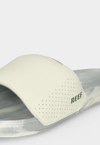 Sandale à enfiler gris clair avec large bride crème comportant de petites perforations et logo vert "REEF" sur le côté.